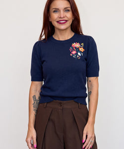 A woman wearing a Bob & Bill navy knit short-sleeve sweater with colorful floral embroidery on the chest.